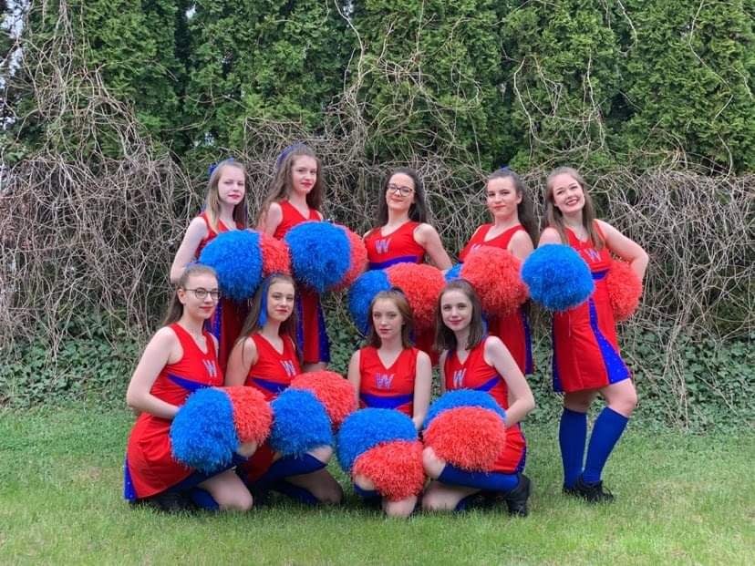 Cheerleaderki  9 dziewcząt ubranych w stroje w barwach   czerwono niebieskich, z pomponami w rękach pozują do zdjęcia